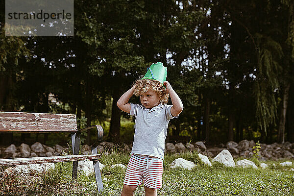 Niedlicher blonder Junge mit Eimer auf dem Kopf  der neben einer Bank im Hinterhof steht