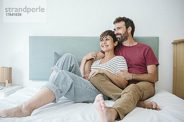Glückliches Paar mittleren Alters schaut weg  während es zu Hause zusammen im Bett sitzt