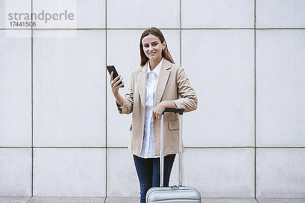 Lächelnde Geschäftsfrau mit Handy und Gepäck vor weißer Wand