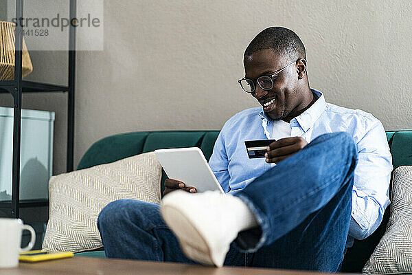 Lächelnder Mann  der mit Kreditkarte bezahlt  während er zu Hause ein digitales Tablet nutzt