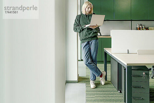 Geschäftsfrau benutzt Laptop  während sie sich im Büro an eine architektonische Säule lehnt