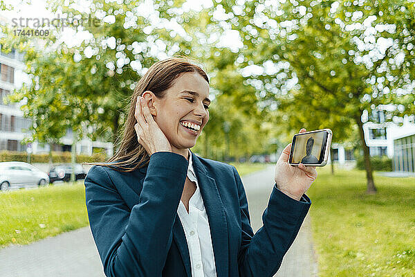 Glückliche Geschäftsfrau mit der Hand im Haar während eines Videoanrufs im öffentlichen Park