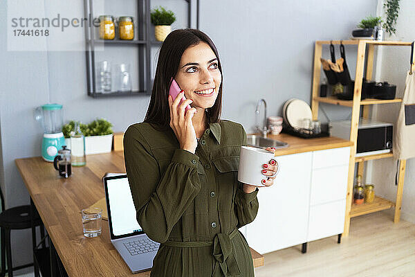 Lächelnde Berufstätige mit Kaffeetasse  die zu Hause mit dem Handy telefoniert