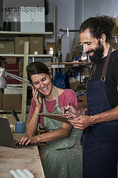 Glückliche Unternehmerin  die mit dem Smartphone spricht  während sie neben einem Mann sitzt  der in der Werkstatt ein Klemmbrett hält