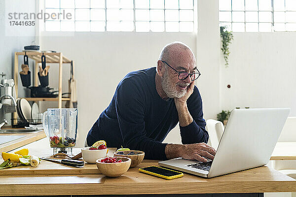 Älterer Mann benutzt Laptop  während er sich an die Küchentheke lehnt