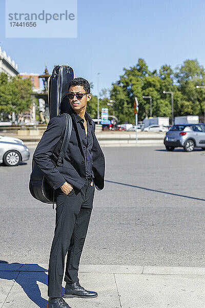 Junger Musiker trägt Gitarrenkoffer  während er an sonnigen Tagen mit den Händen in den Taschen auf der Straße steht