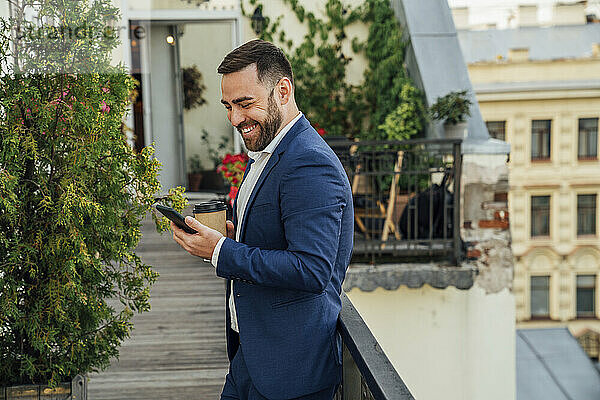 Lächelnder Geschäftsmann benutzt Smartphone in der Nähe des Geländers auf dem Bürobalkon