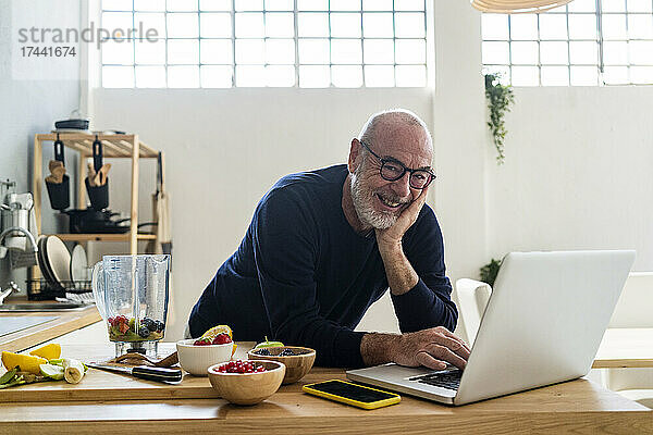 Lächelnder Mann mit Laptop lehnt an der Küchentheke