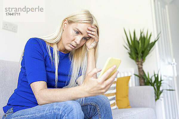 Besorgte Frau mit Kopf in der Hand  die ihr Smartphone hält  während sie zu Hause sitzt