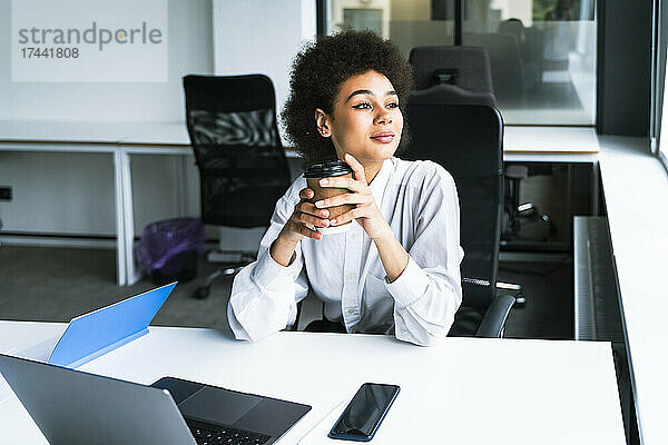 Geschäftsfrau hält Einwegkaffeetasse in der Hand  während sie mit Laptop im Büro sitzt