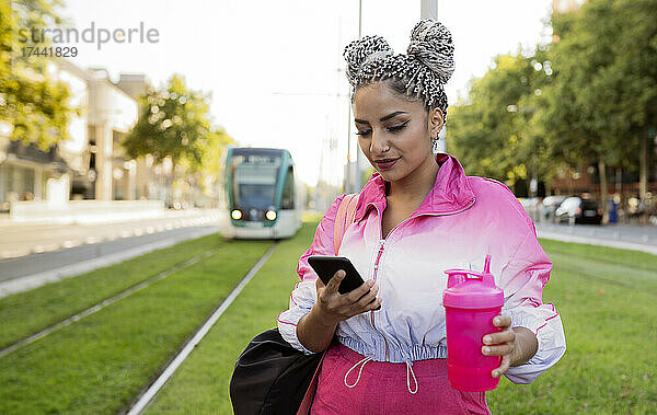 Frau mit Wasserflasche benutzt Smartphone an Straßenbahngleis