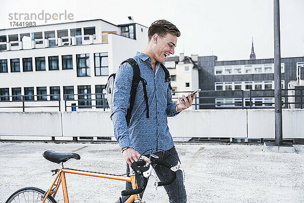 Fröhlicher Mann mit Fahrrad und Handy beim Gehen auf dem Dach des Parkhauses
