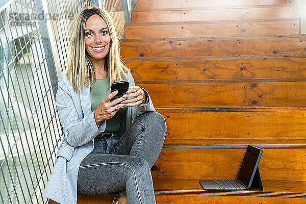 Blonde Frau mit Mobiltelefon und digitalem Tablet sitzt auf Stufen