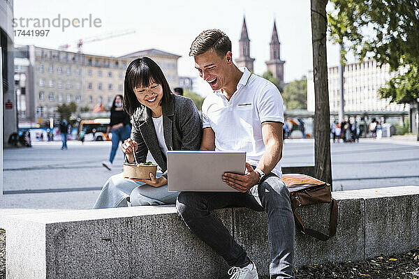 Glückliche männliche und weibliche Freunde  die Laptop benutzen  während sie auf der Stützmauer sitzen