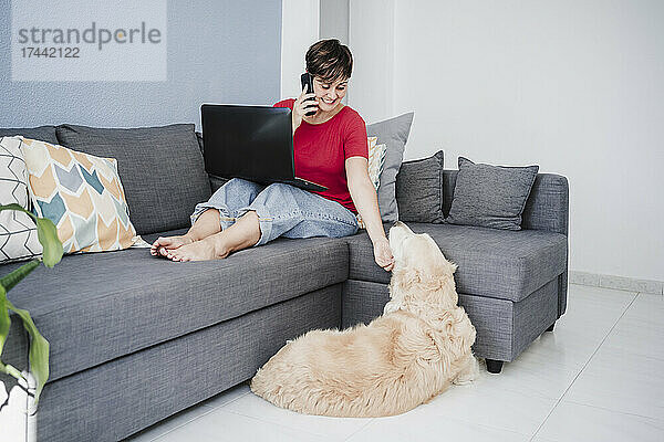 Geschäftsfrau mit Laptop streichelt Hund  während sie zu Hause mit dem Smartphone spricht