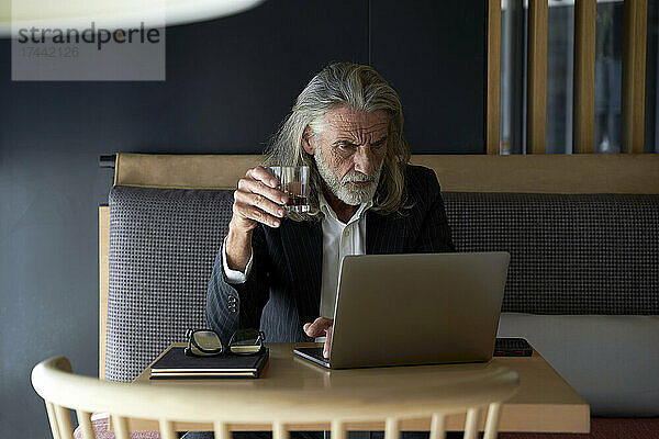 Männlicher Geschäftsmann hält Whiskeyglas in der Hand  während er im Hotelcafé am Laptop arbeitet