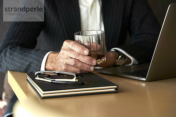 Reifer Geschäftsmann hält Whiskeyglas am Laptop im Hotel