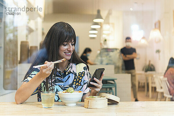 Lächelnde Frau mittleren Alters benutzt Smartphone  während sie im Restaurant Poke isst