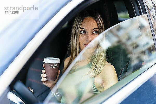 Blonde Frau mit Kaffeetasse schaut durch das Autofenster