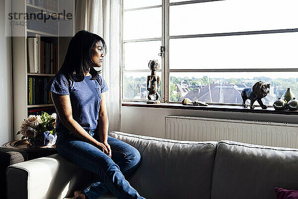 Frau schaut durchs Fenster  während sie zu Hause auf dem Sofa sitzt