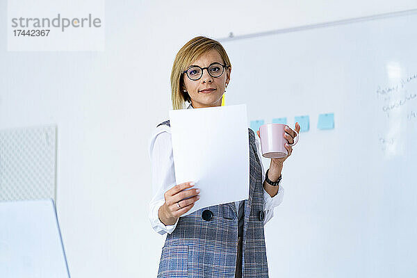 Geschäftsfrau mit Dokument und Kaffeetasse steht am Whiteboard im Büro