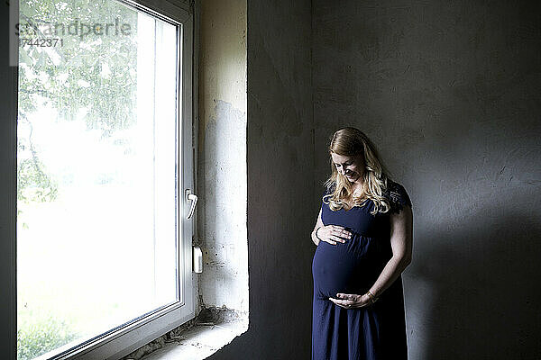 Schwangere Frau berührt Bauch  während sie am Fenster steht