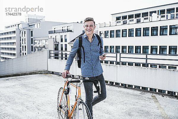 Lächelnder Mann mit Smartphone  der Fahrrad fährt  während er auf dem Dach läuft