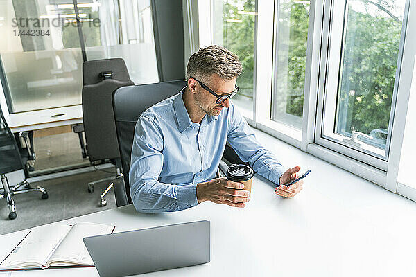 Geschäftsmann mit Kaffeetasse und Mobiltelefon am Schreibtisch im Büro