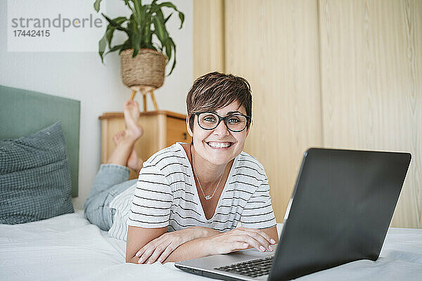 Lächelnde Frau mittleren Alters mit Laptop auf dem Bett liegend