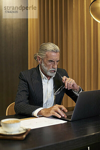 Reifer Geschäftsmann hält eine Brille in der Hand  während er im Hotel am Laptop arbeitet