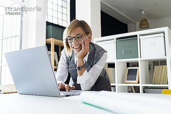 Lächelnde Geschäftsfrau mit der Hand am Kinn  die einen Laptop benutzt  während sie sich auf den Schreibtisch lehnt