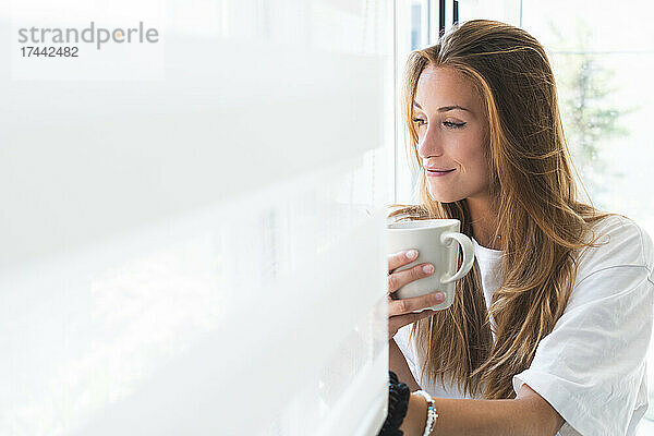 Schöne junge Frau mit Tasse  die zu Hause durch das Fenster schaut