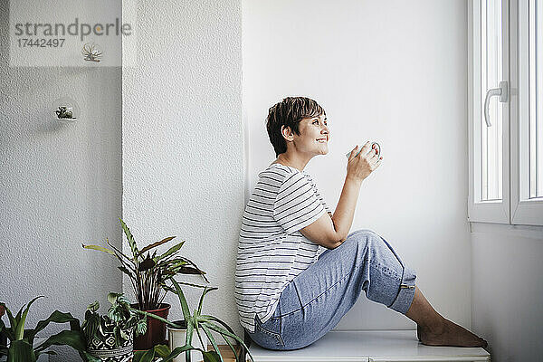 Lächelnde Frau mittleren Alters hält einen Becher in der Hand  während sie zu Hause neben Pflanzen sitzt