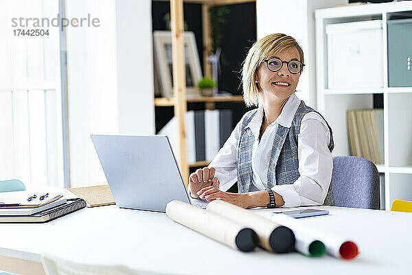 Lächelnde Geschäftsfrau mit Laptop sitzt am Schreibtisch im Büro