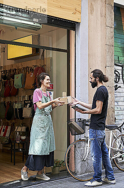 Lächelnde Unternehmerin gibt dem männlichen Zusteller Paket  während sie vor dem Geschäft steht