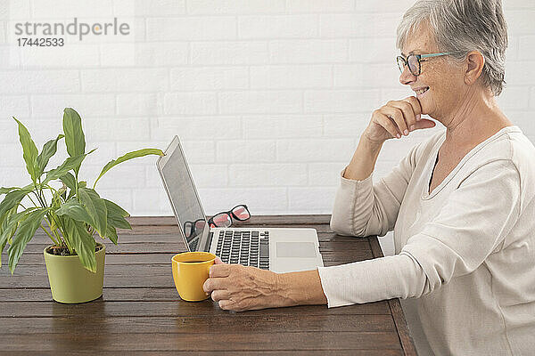 Lächelnde Geschäftsfrau beim Kaffeetrinken während der Videokonferenz zu Hause