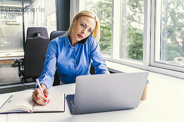 Blonde Geschäftsfrau benutzt Laptop  während sie im Büro mit dem Smartphone spricht