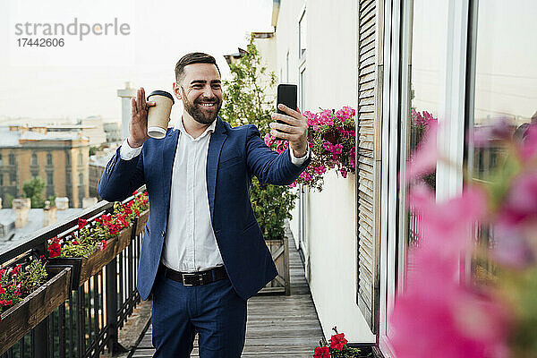 Lächelnder Geschäftsmann winkt während eines Videoanrufs auf dem Smartphone auf dem Balkon