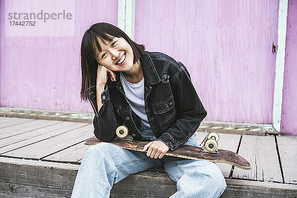 Glückliches Teenager-Mädchen mit Skateboard sitzt vor der Wand