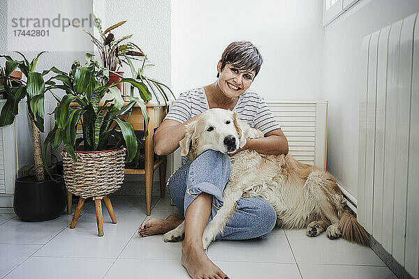 Lächelnde Frau sitzt mit Hund zu Hause neben Pflanzen