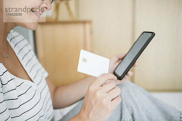 Frau hält Kreditkarte in der Hand  während sie zu Hause ihr Smartphone benutzt