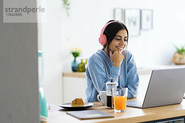 Lächelnde junge Frau mit der Hand am Kinn während eines Videoanrufs über den Laptop zu Hause