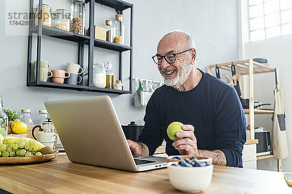 Glücklicher älterer Mann hält Apfel in der Hand  während er zu Hause Laptop benutzt