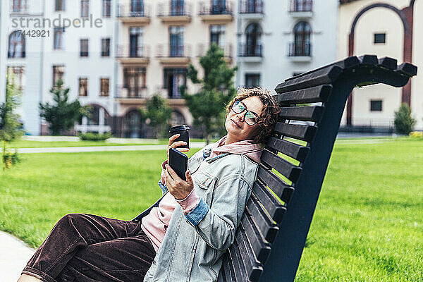 Frau mittleren Alters mit Mobiltelefon trinkt Kaffee  während sie auf einer Bank sitzt