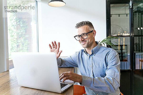 Lächelnder Geschäftsmann winkt während eines Videoanrufs über den Laptop im Büro