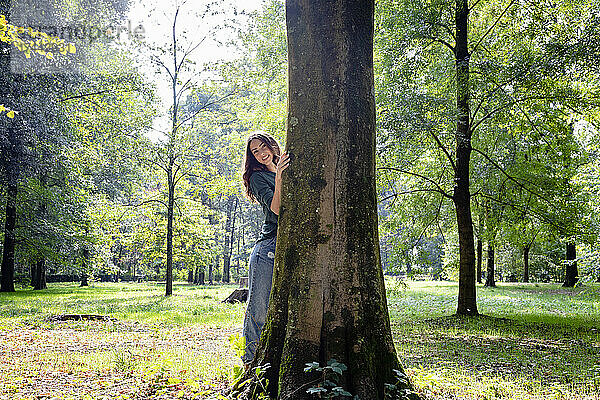 Lächelnde Frau steht hinter Baumstamm im Park