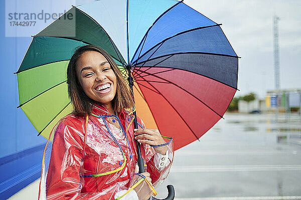 Glückliche Frau im Regenmantel mit buntem Regenschirm während der Regenzeit