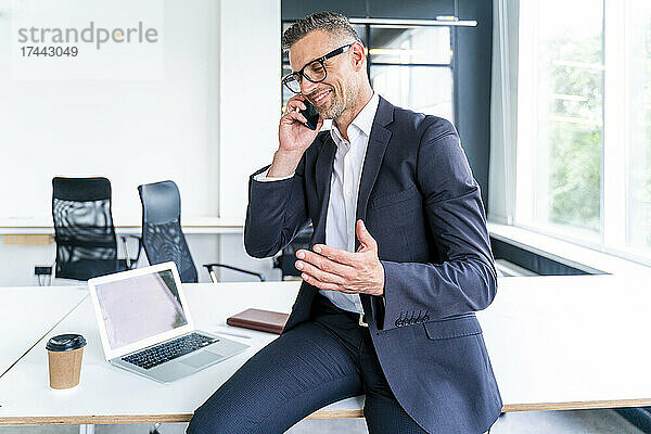 Lächelnder Geschäftsmann gestikuliert  während er im Büro mit dem Mobiltelefon spricht