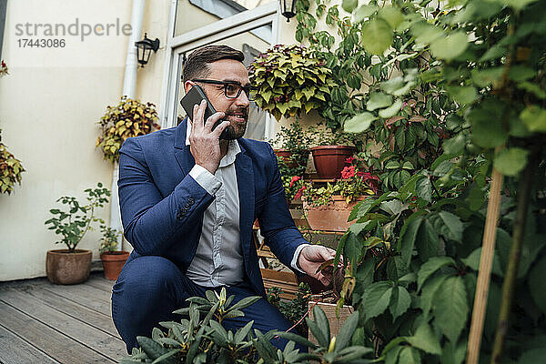 Geschäftsmann telefoniert  während er neben Pflanzen auf dem Balkon hockt