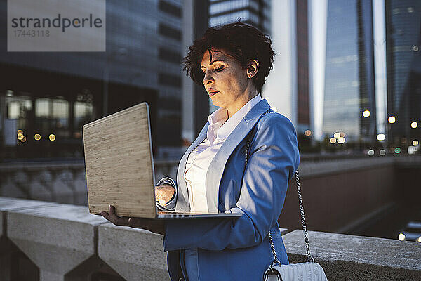 Berufstätige Frau benutzt Laptop  während sie in der Abenddämmerung am Geländer der Brücke steht